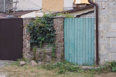 part of a fence wall made of brown stones and wooden boards overgrown with green vegetation on a rural street