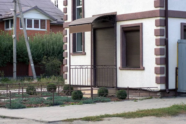 gray wall of the house with a window and door covered with brown roller blinds in the street