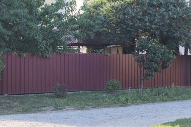 Kırsal bir sokakta yeşil çimenler ve bitki örtüsü içinde uzun kahverengi metal çitler.