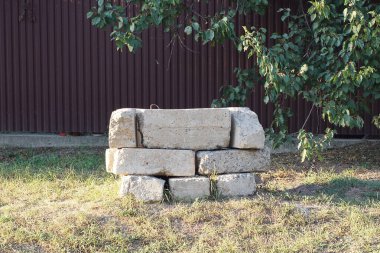 Çitin kahverengi duvarının yanındaki yeşil çimlerde, caddede gri beton bloklar yığını var.