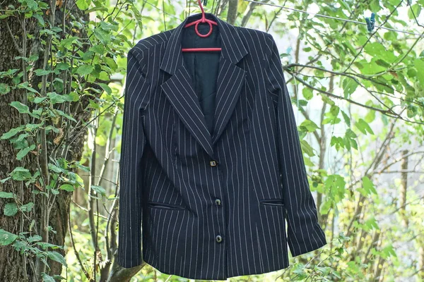 one black striped old jacket made of fabric hangs on a red hanger and a wire in the street among green vegetation