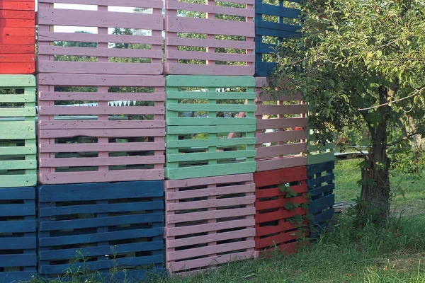 a large colored wall of wooden pallets in green grass near a tree on the street