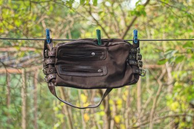 Deri ve kumaştan yapılmış kahverengi bir çanta yeşil bitkilerin arasında bir tele asılmış.