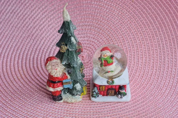 一套圣诞玩具 配有绿树的桑塔爪和粉色桌子上的玻璃球里的雪人 — 图库照片