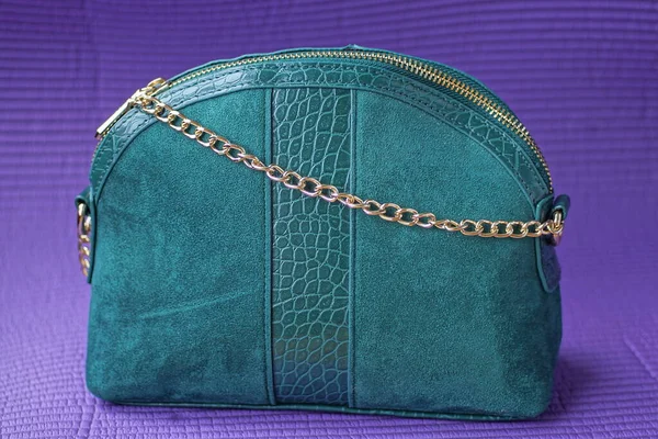 one green bag made of suede and leather with a yellow metal chain and harness stands on a purple table