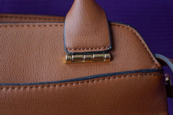 part of a brown leather bag with a handle with a metallic yellow fastener on a purple background
