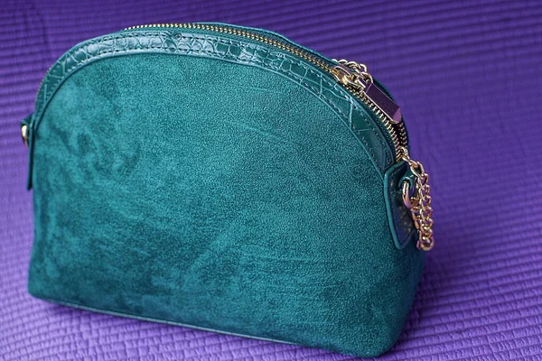 one green bag made of suede and leather with a yellow metal chain and harness stands on a purple table