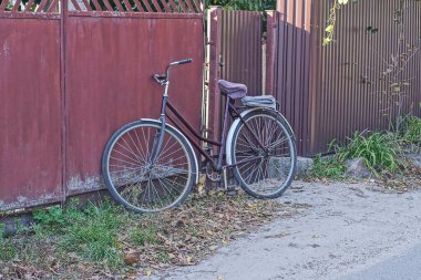 Eski bir bisiklet gri asfalt bir yolun yanındaki kırsal bir sokakta kahverengi bir demir çitin karşısında duruyor.
