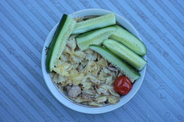 Bir tabak dolusu yeşil salatalıklı makarna ve gri bir masada duran kırmızı domates.