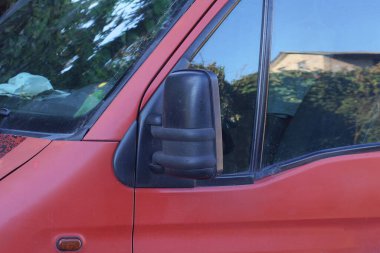 Caddedeki kırmızı bir arabanın gri camının yanında büyük siyah plastik bir araba aynası.