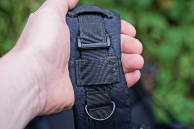 El, yeşil bir arka planda taktik sırt çantasında metal bir halkayla büyük bir kumaş kayışı tutuyor.