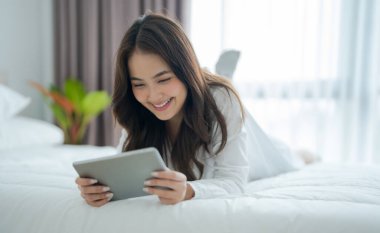 Yatakta uzanmış, yatak odasında tablet bilgisayarla oynuyor. Gülümseyen bir yüzü var, mutlu ve rahatlamış bir ifadesi var..