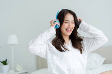 Beyaz yatak odasında yatağında oturan bir kadın müzik dinler, müziğin tadını çıkarır, gülümser, ferahlar ve dinlenir..