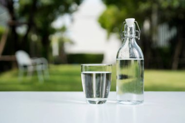 Bir bardak temiz içme suyu ve bir su şişesi beyaz ahşap bir masanın üzerinde doğal bir bahçe arka planına sahip..