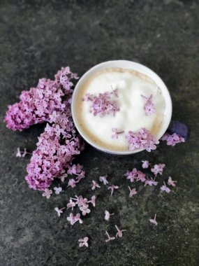 Siyah taş bir masada bir fincan kahvenin içinde pembe leylak çiçeği..