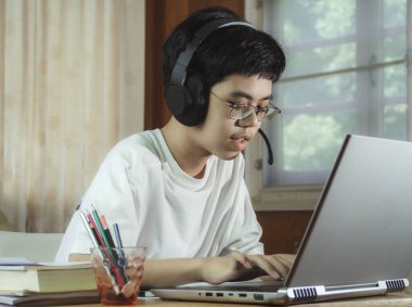 Kulaklık takan çocuk internette sohbet eden Skype öğretmeniyle masada oturuyor sınava hazırlanıyor ve ödevi için dizüstü bilgisayar kullanıyor ve online eğitim için E-öğrenim görüyor. çocuk konsepti öğrenmek.