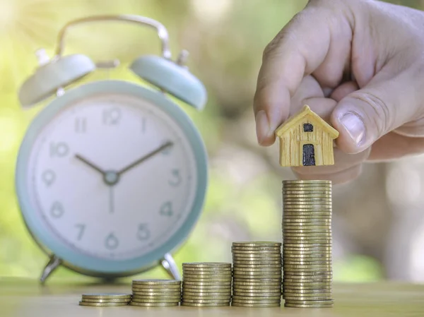 Hand are stacking coins and home with alarm clock to save the financial stability for the future. Concept for loan, property ladder, financial, mortgage, real estate investment, taxes and bonus.