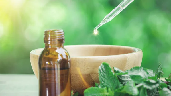 The peppermint extract in small bottle near peppermint leaf on wooden table. The essential oil falling from glass dropper into organic bio alternative medicine, brown bottle.