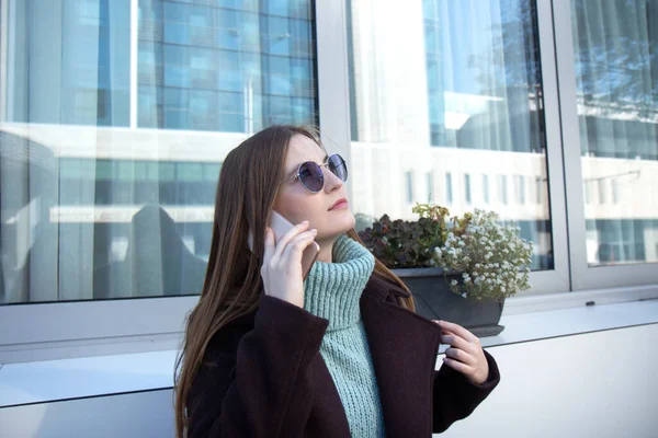 Mulher bonita com cabelos longos em um casaco escuro caminha pela rua e fala em um telefone celular — Fotografia de Stock