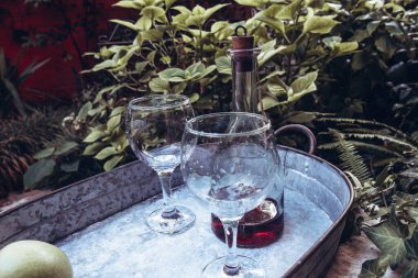 Bir kadeh şarap, iki bardak ve bahçede büyük bir demir tepside olgun yeşil elmalar. Yan görünüm