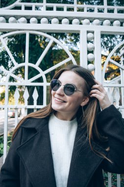 Gülümseyen, gözlüklü, uzun saçlı, siyah ceketli güzel bir kadın sokakta yürüyor.