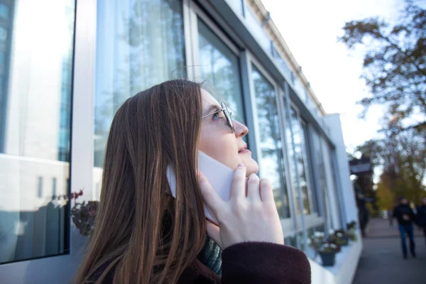 Žena s dlouhými vlasy v tmavém kabátě kráčí ulicí a mluví na mobilním telefonu — Stock fotografie