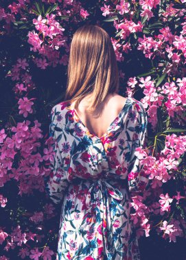 Sırtı güzel pembe çiçeklerle dolu genç bir kadın. Çiçekler arasında renkli keten elbiseli bir kız. Arkadan bak. Dikey fotoğraf
