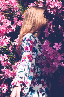 Sırtı güzel pembe çiçeklerle dolu genç bir kadın. Çiçekler arasında renkli keten elbiseli bir kız. Arkadan bak. Dikey fotoğraf