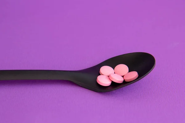 Vitamin pink tablets on black spoon on a violet background. Dietary supplements. Complementary and medical products