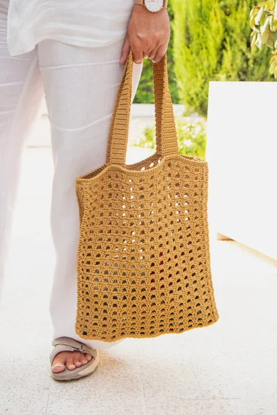 Woman holds a handmade beige knitted bag in her hand near her legs outdoors. Sustainable shopping. Wasteless lifestyle. Female with a jute bag with her own hands on a walk. Close up