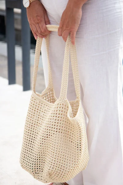 Woman holds a handmade beige knitted bag in her hand near her legs outdoors. Sustainable shopping. Wasteless lifestyle. Female with a jute bag with her own hands on a walk. Close up