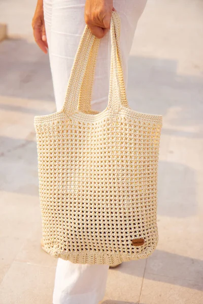 Woman holds a handmade beige knitted bag in her hand near her legs outdoors. Sustainable shopping. Wasteless lifestyle. Female with a jute bag with her own hands on a walk. Close up