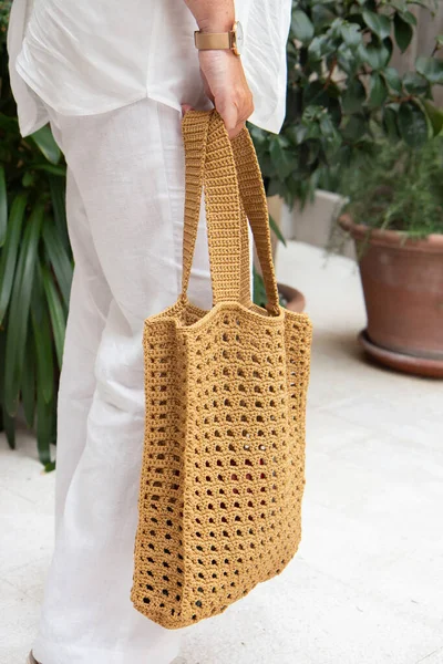 Woman holds a handmade beige knitted bag in her hand near her legs outdoors. Sustainable shopping. Wasteless lifestyle. Female with a jute bag with her own hands on a walk. Close up