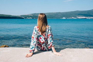 Güzel kız sırtı denize dönük oturuyor. Parlak elbiseli, uzun sarı saçlı bir kadın. Yaz sahili. Arka plan