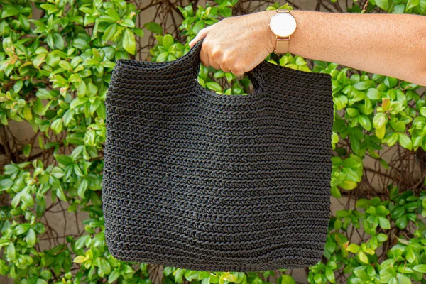 Woman holds a handmade black knitted bag in her hand outdoors. Sustainable shopping. Wasteless lifestyle. Female with a jute bag with her own hands on a walk. Close up
