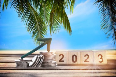 New year 2023 on wooden cube and stack of coins with arrow upwards on planks on tropical beach background. Business beginning with success and return on investment idea