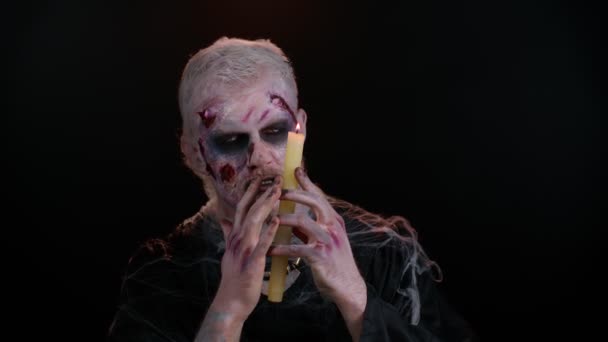 Finsterer Mann mit furchteinflößendem Halloween-Zombie-Make-up, das Gesichter macht, zaubert Zaubersprüche über eine Kerze — Stockvideo