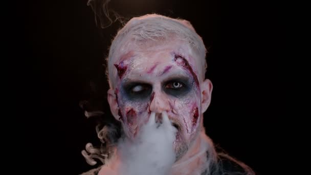 Zombie hombre con maquillaje con cicatrices heridas y lentes de contacto blancos sopla humo de la nariz y la boca — Vídeos de Stock
