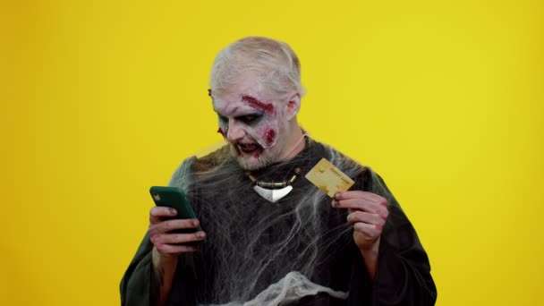 Чоловік-синістер Хеллоуїн зомбі за допомогою кредитної банківської картки та смартфона під час покупки в Інтернеті — стокове відео