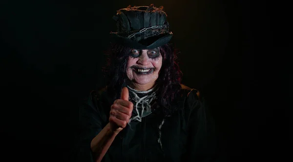 Зловещая пожилая женщина со страшным макияжем ведьмы Хэллоуина в костюме поднимает большие пальцы вверх, как жест — стоковое фото