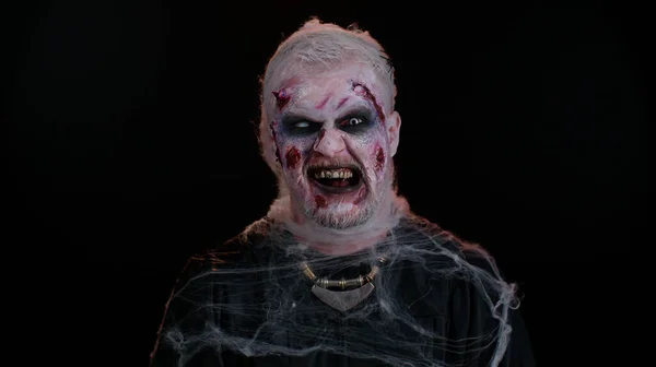 Зомби-человек со шрамами от ран и контактными линзами, глядя в камеру, щёлкает зубами, пытаясь напугать — стоковое фото
