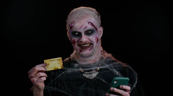 Чоловік-синістер Хеллоуїн зомбі за допомогою кредитної банківської картки та смартфона під час покупки в Інтернеті — стокове фото
