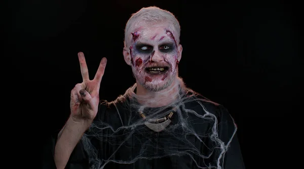 Halloween zombie man bloody make-up showing victory sign, hoping for success and win peace gesture