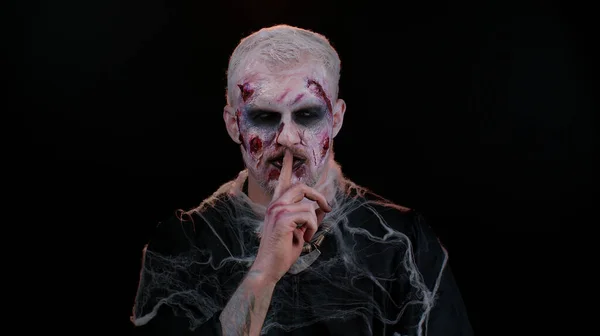 Halloween zombie man presses index finger to lips makes silence gesture sign do not tells secret