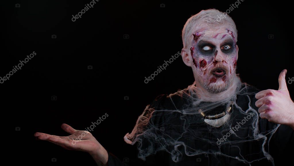 Zombie man with makeup with fake wounds scars showing thumbs up and pointing empty place, advertising area for commercial text logo, copy-space for goods promotion. Sinister Halloween ugly dead guy