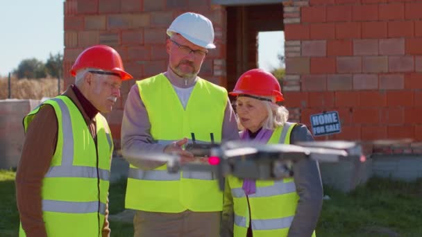 Trabajador especialista vuela en dron en el sitio de construcción de edificios, mostrando la casa a los abuelos clientes — Vídeos de Stock