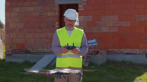 Operador de drones con mando a distancia controla una aeronave frente al sitio de la casa de construcción — Vídeo de stock