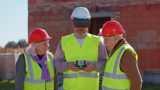 Trabajador especialista vuela en dron en el sitio de construcción de edificios, mostrando la casa a los abuelos clientes — Vídeos de Stock