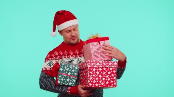 Hombre encantador en Navidad suéter rojo sombrero presentando cajas de regalo de Navidad se extiende la mano a la cámara — Vídeos de Stock