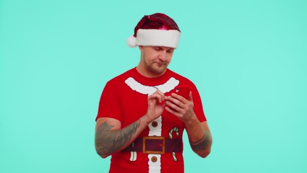 Homem de t-shirt de Natal, chapéu usando telefone celular digitando novo post na web, compras on-line, navegação — Vídeo de Stock